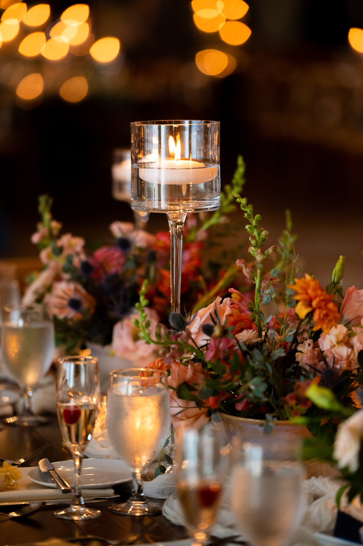 Elegant table setting at Hotel LBI Ship Bottom NJ with floral arrangements, lit candle in glass holder, and filled wine glasses. Blurred bokeh lights create a warm, inviting atmosphere, perfect for capturing New Jersey's finest Jersey Shore wedding venues.