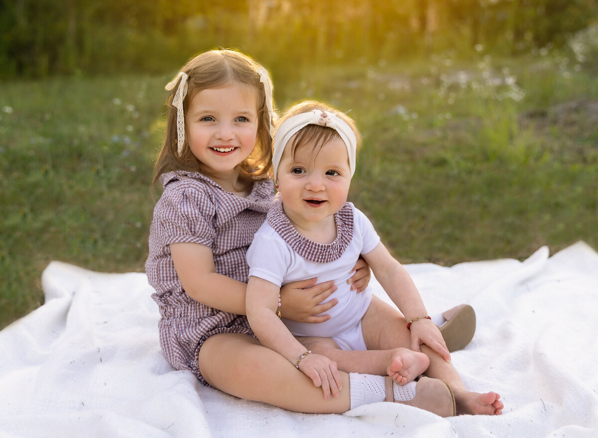 Two sisters sit for a family photoshoot in a brooklyn, ny park at golden hour. Both girls are sitting on a blanket with big sister, approx age 4 holding her little sister, approx. age 1. Both of the girls are smiling at the camera and are surrounded by soft golden light. Captured by premier Brooklyn NY family photographer Chaya Bornstein Photography.