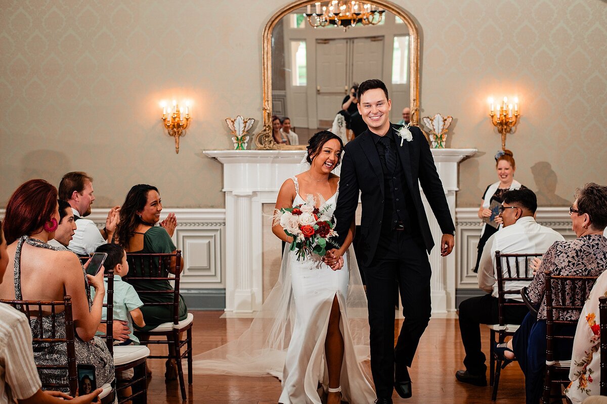 The groom, wearing a black suit with a black shirt and tie holds hands with the bride wearing a white gown with a thigh high slit and spaghetti straps as they walk down the aisle at Maney Hall to cheers. The bride is holding a boho bouquet of crimson dahlias, orange roses, silver dollar eucalyptus, ivory roses and pampas grass. Behind them is a white mantle and large fold framed mirror.