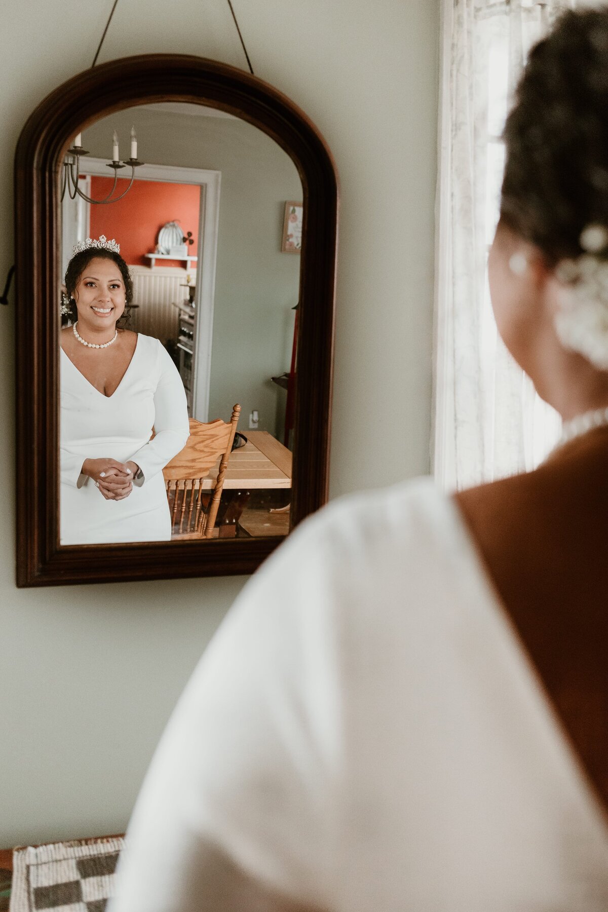 Bride in a white gown with a tiara smiles as she looks at her reflection in a vintage arched mirror, with the cozy interior of a room subtly reflected behind her.