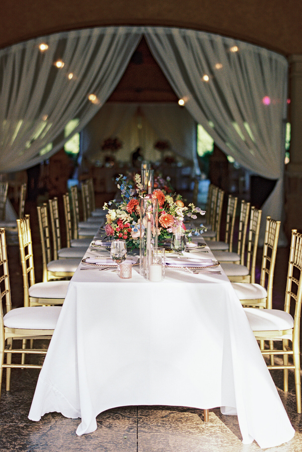 Long reception table with gold chairs, colorful floral centerpieces, and white draping at Gervasi Vineyard wedding.