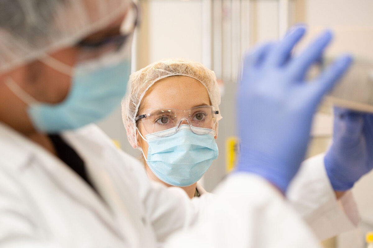 Scientist wearing protective glasses, a face mask and a hair net looking at a tray held by another out of focus scientist