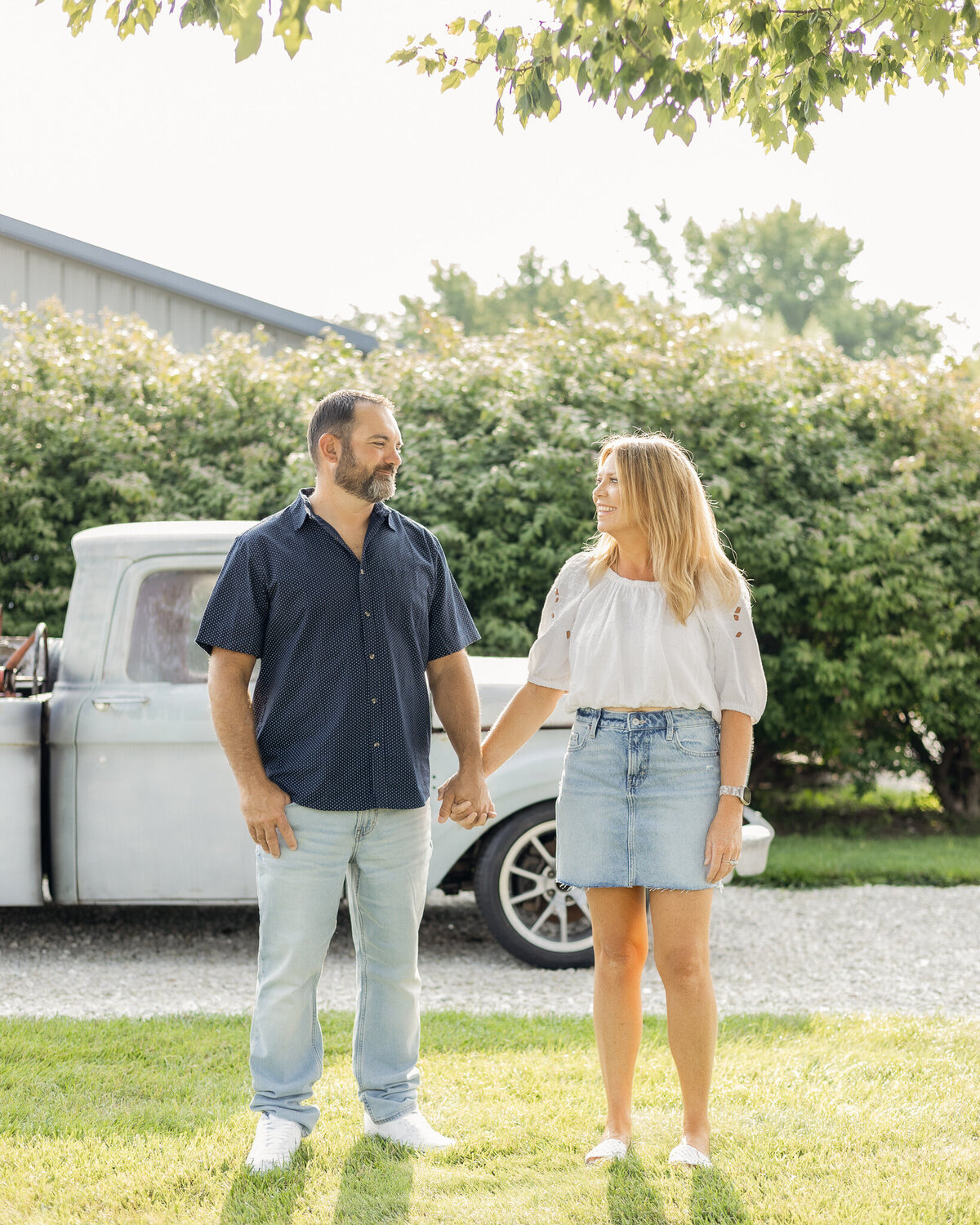 Husband and wife standing next to each other in grass with a classic truck behind them.  They are looking at each other smiling.  Wife is wearing a jean skirt, white half sleeve shirt, and has long blonde hair.  Husband is wearing a blue button up short sleeve shirt, blue jeans, and has black gray short hair and beard.