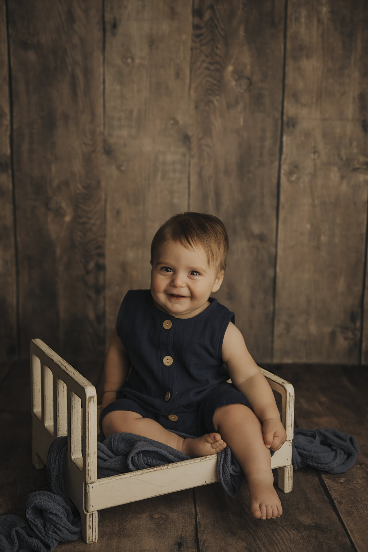 boy on white bed in blue outfit on wood backdrop in milestone session