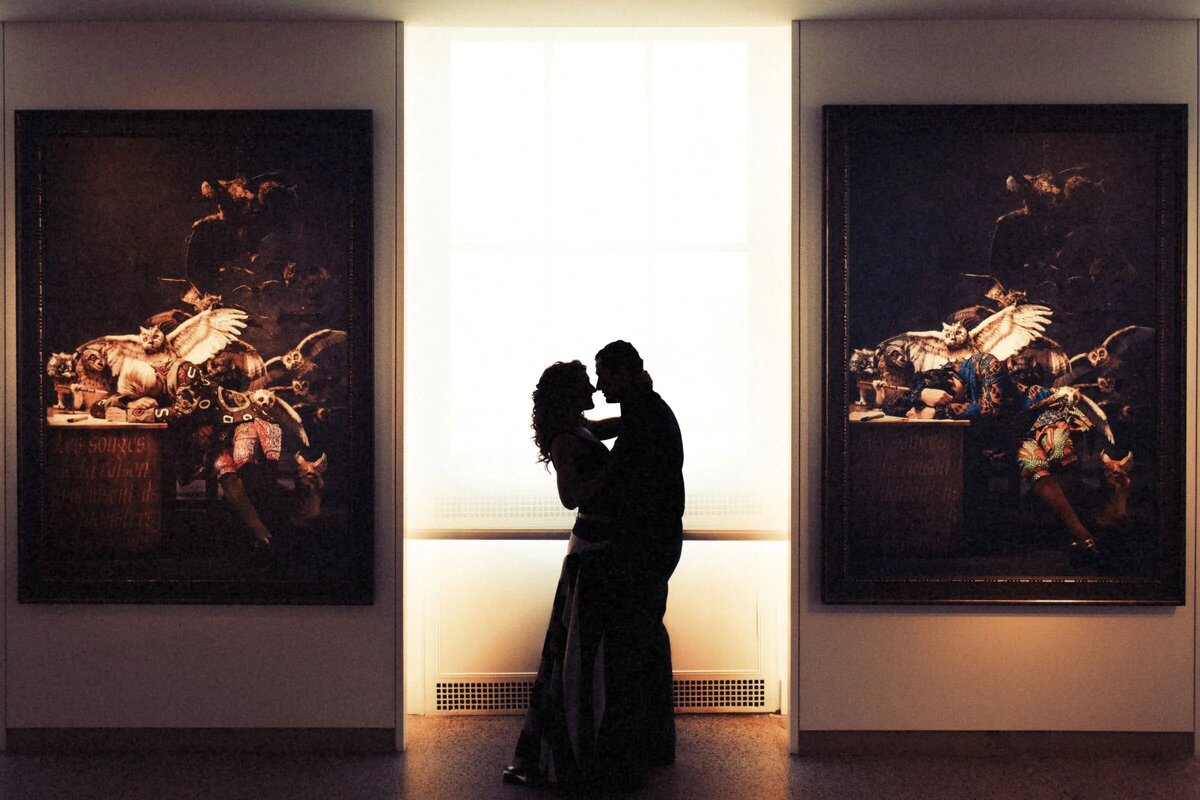 Silhouette of a couple kissing in an art gallery, framed between two large, dark paintings, with a soft light from behind