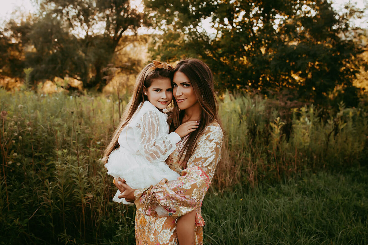 a mother holding her little girl with their heads close looking at the camera in a gorgeous outdoor setting