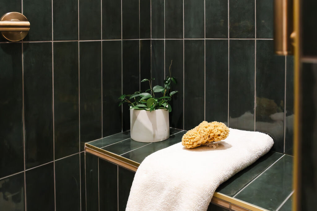 A detail of a shower area with dark green vertical tiles and light grout lines. A built-in tiled bench with a gold trim holds a rolled-up white towel, a natural sea sponge, and a small white pot containing a trailing green plant, possibly a Hoya. A brushed gold shower fixture is partially visible on the left wall. The lighting is bright and focused on the bench area.