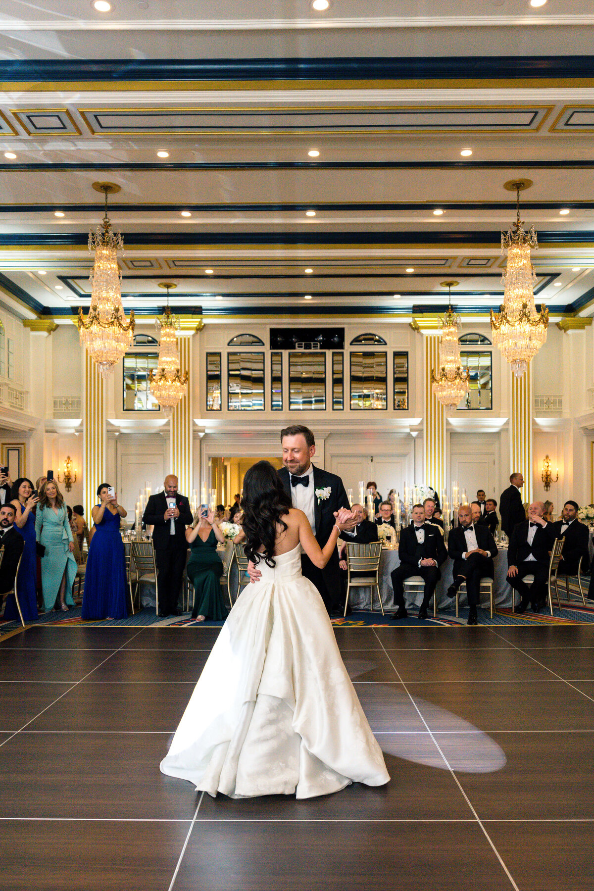 A couple dances in an elegant ballroom adorned with chandeliers and ornate mirrors. The bride wears a flowing white gown, and the groom is in a tuxedo. Guests seated around the room watch and cheer, capturing the moment with phones.