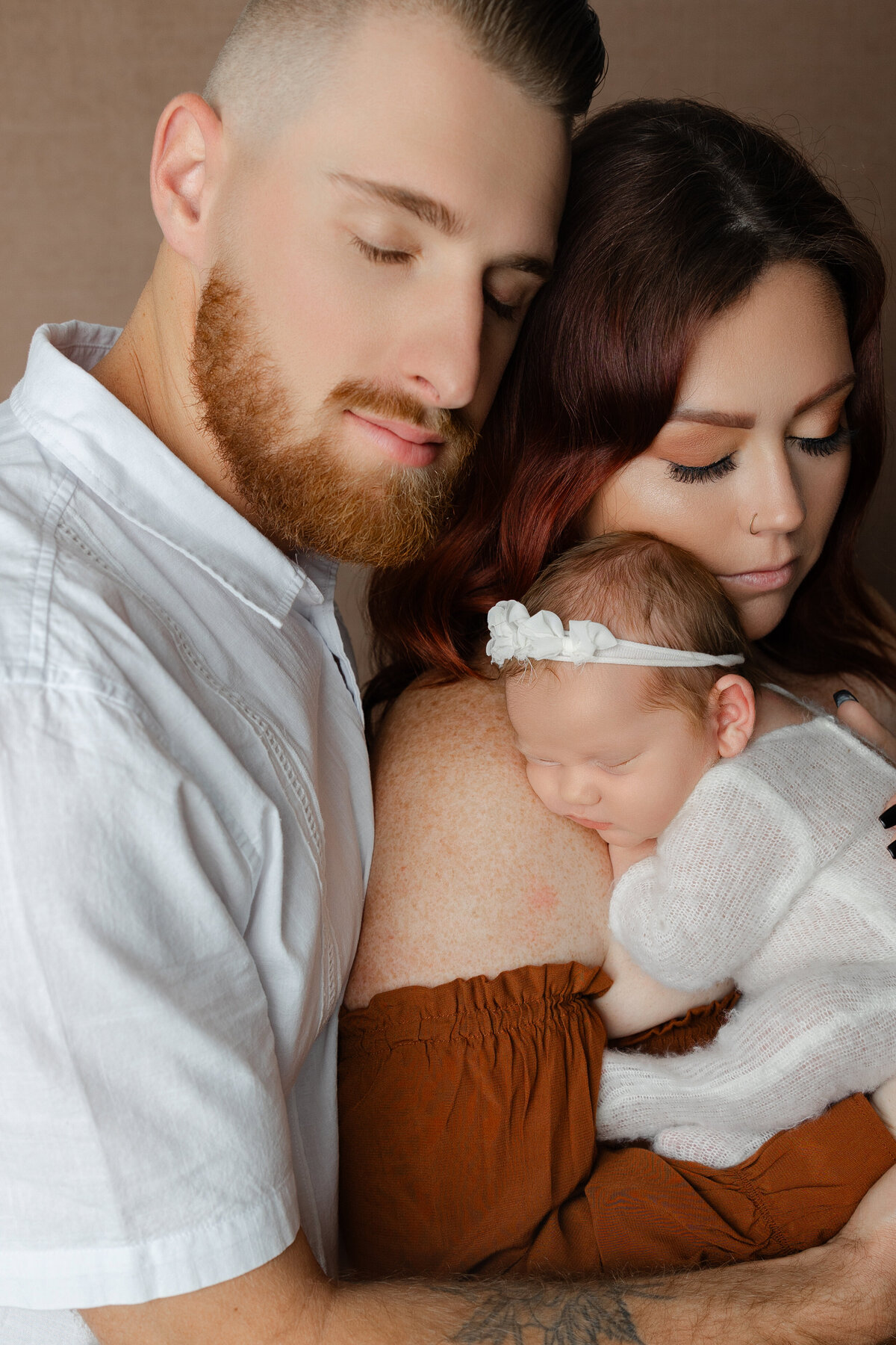 Mom and dad holding their newborn daughter close, with baby swaddled in white and mom gently resting her cheek against her. Photograph by Inland Empire newborn photographer Kaitlyn Dawn Photography.