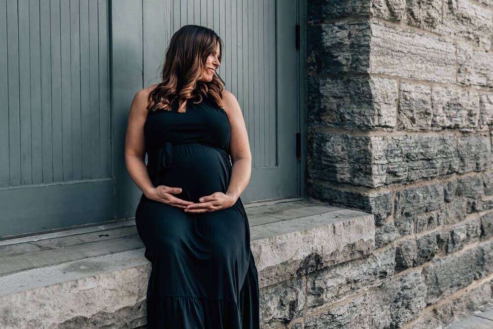 A stunning expecting mother sits framed in the doorway of a stone building with teal doors in the historic St. Anthony Main area of Minneapolis.