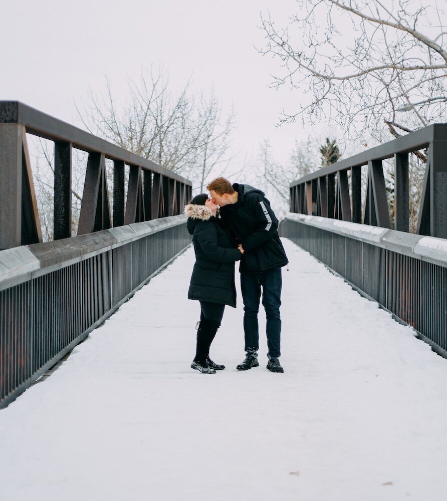 An Edmonton couple kiss on a bridge covered in snow in their black coats