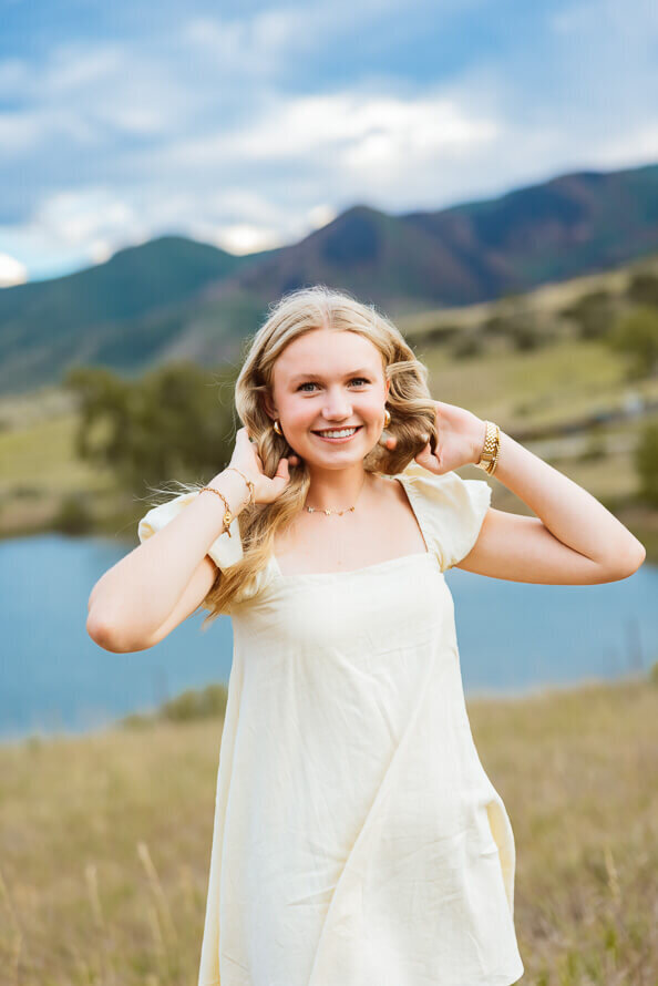 Girl fluffs hair in front of lake and mountains for her senior photos.