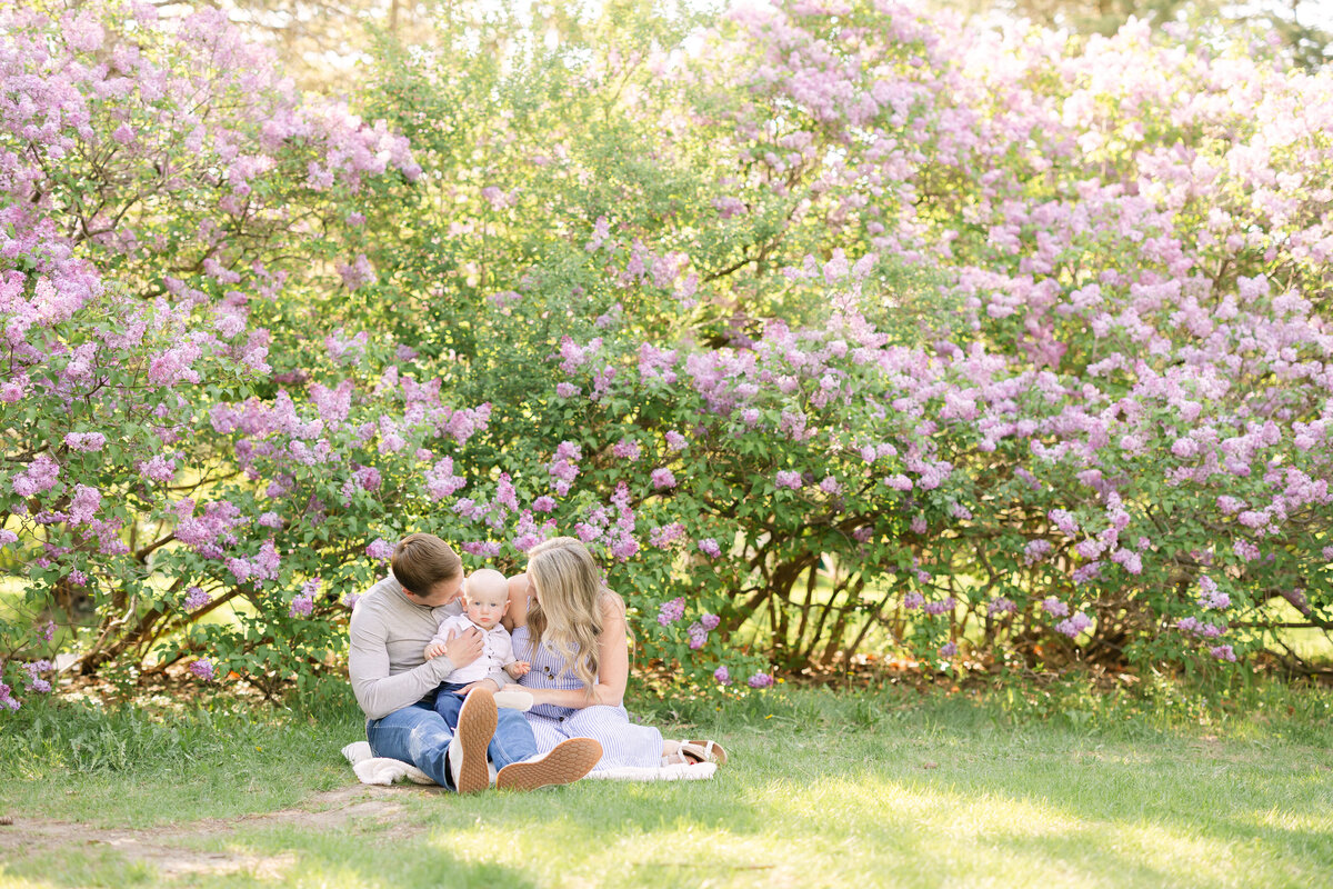a young family sits on a white blanket as they embrace their baby in front of a purple lilac scenery on a sunny spring day