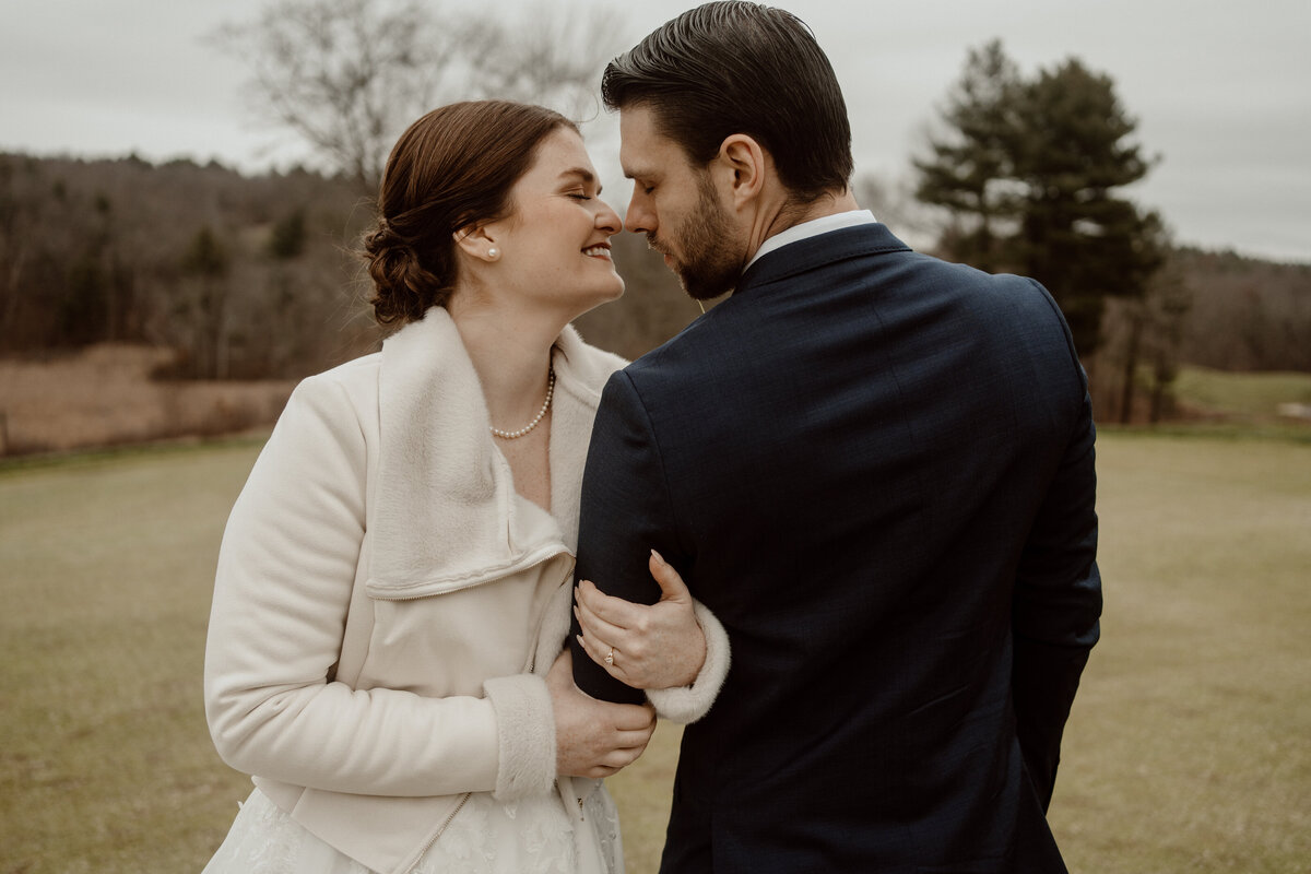 A bride and groom share a tender moment on their wedding day. The bride, wearing a cozy cream jacket over her wedding dress and a string of pearls, lovingly holds onto the groom's arm. The groom, dressed in a navy blue suit, gently leans his forehead against hers. The couple is outdoors, with a serene backdrop of trees and a grassy field.