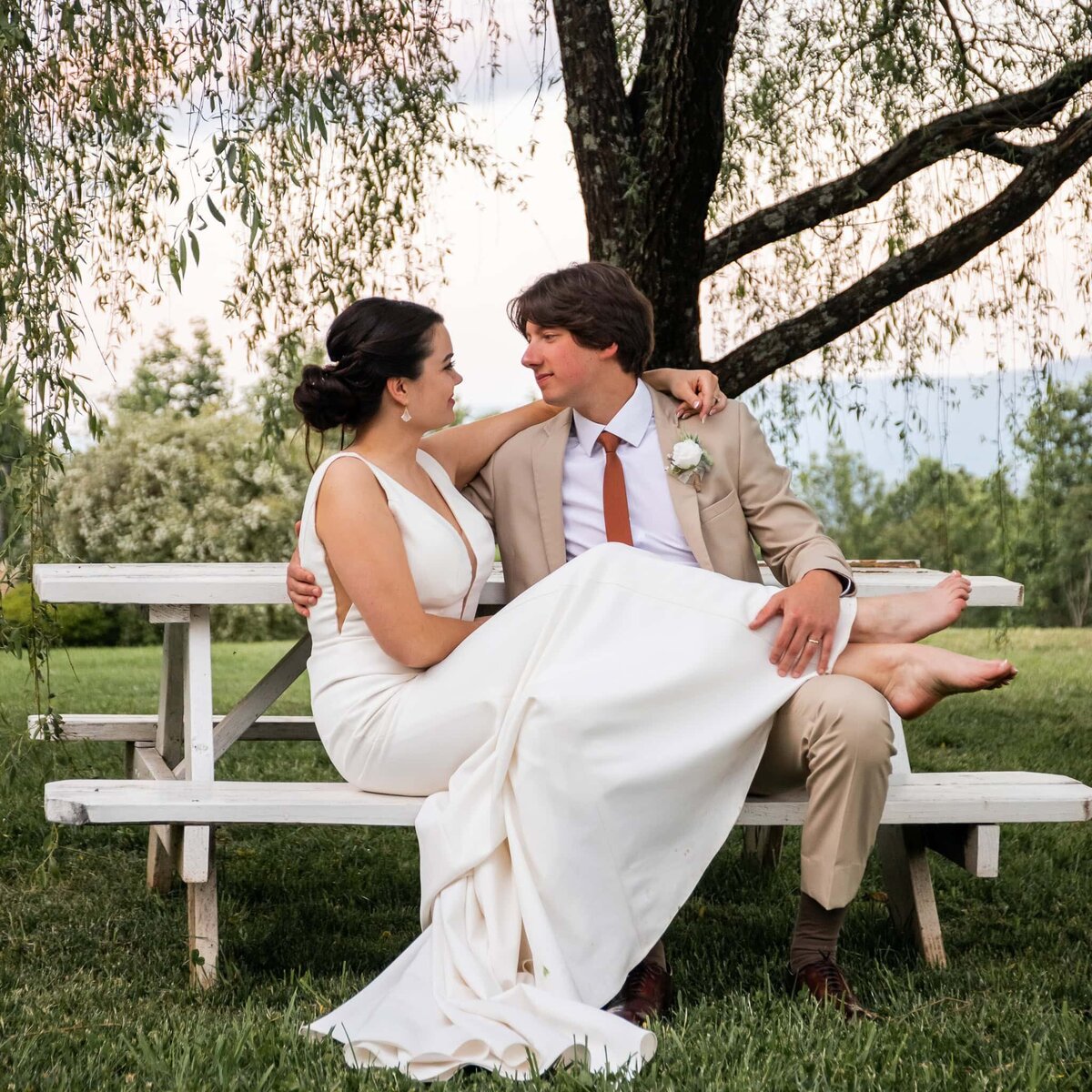 bride and groom sit under a weeping willow tree at on a white picnic table, the bride has thrown her legs over the grooms lap, exposing her bare feet, they both have their arms around each other as they look into each others eyes
