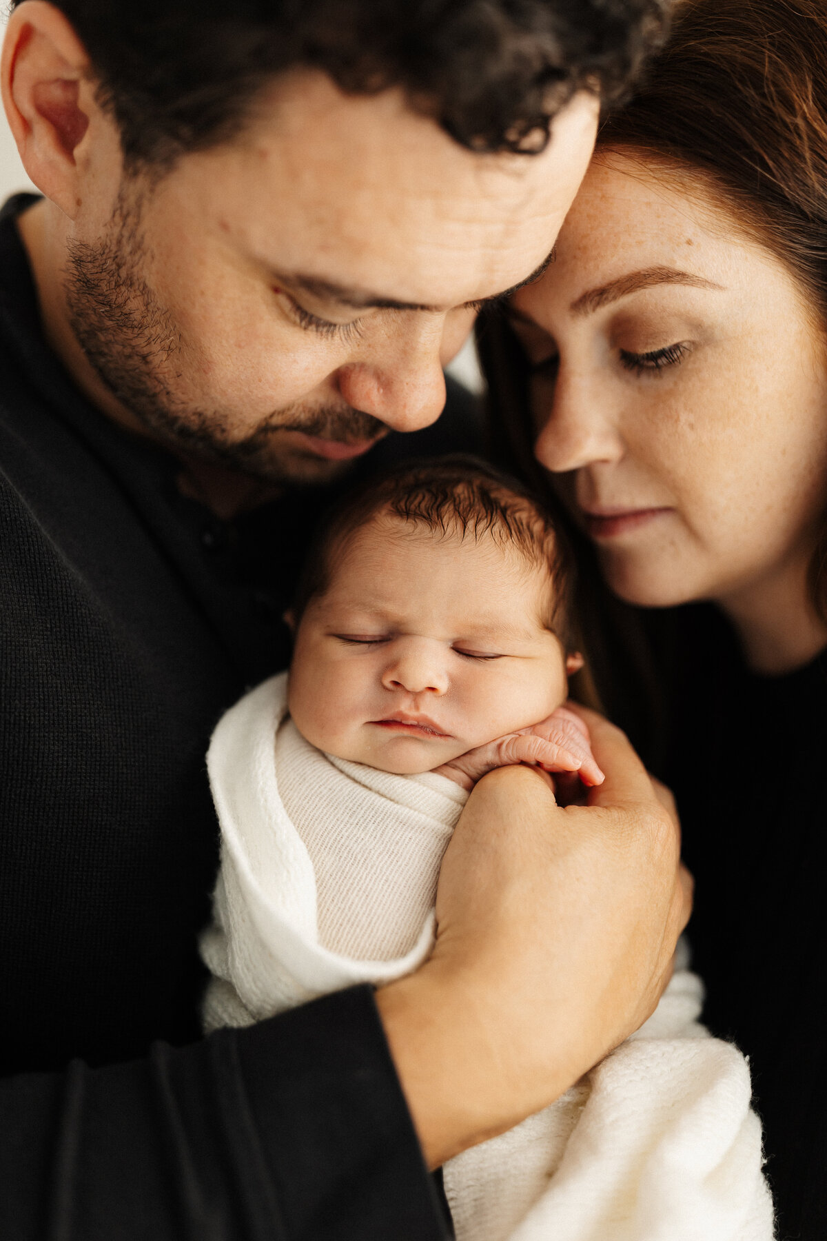 A newborn baby being held by his mother and father while wrapped in a white blanket while his parents pose for their christchurch photographer lucy