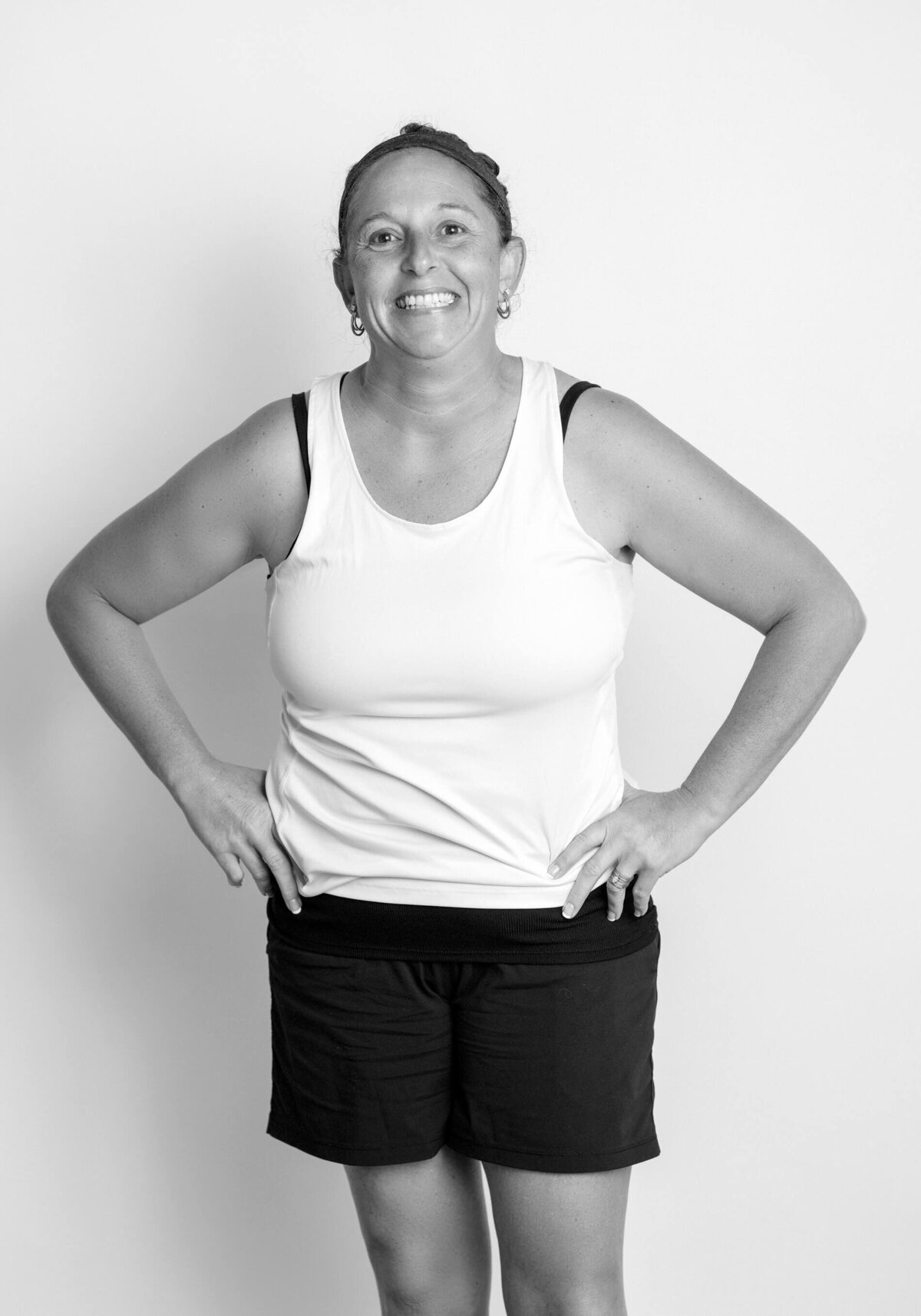 Black-and-white before shot of a woman in a tank top and shorts, smiling confidently with hands on hips.