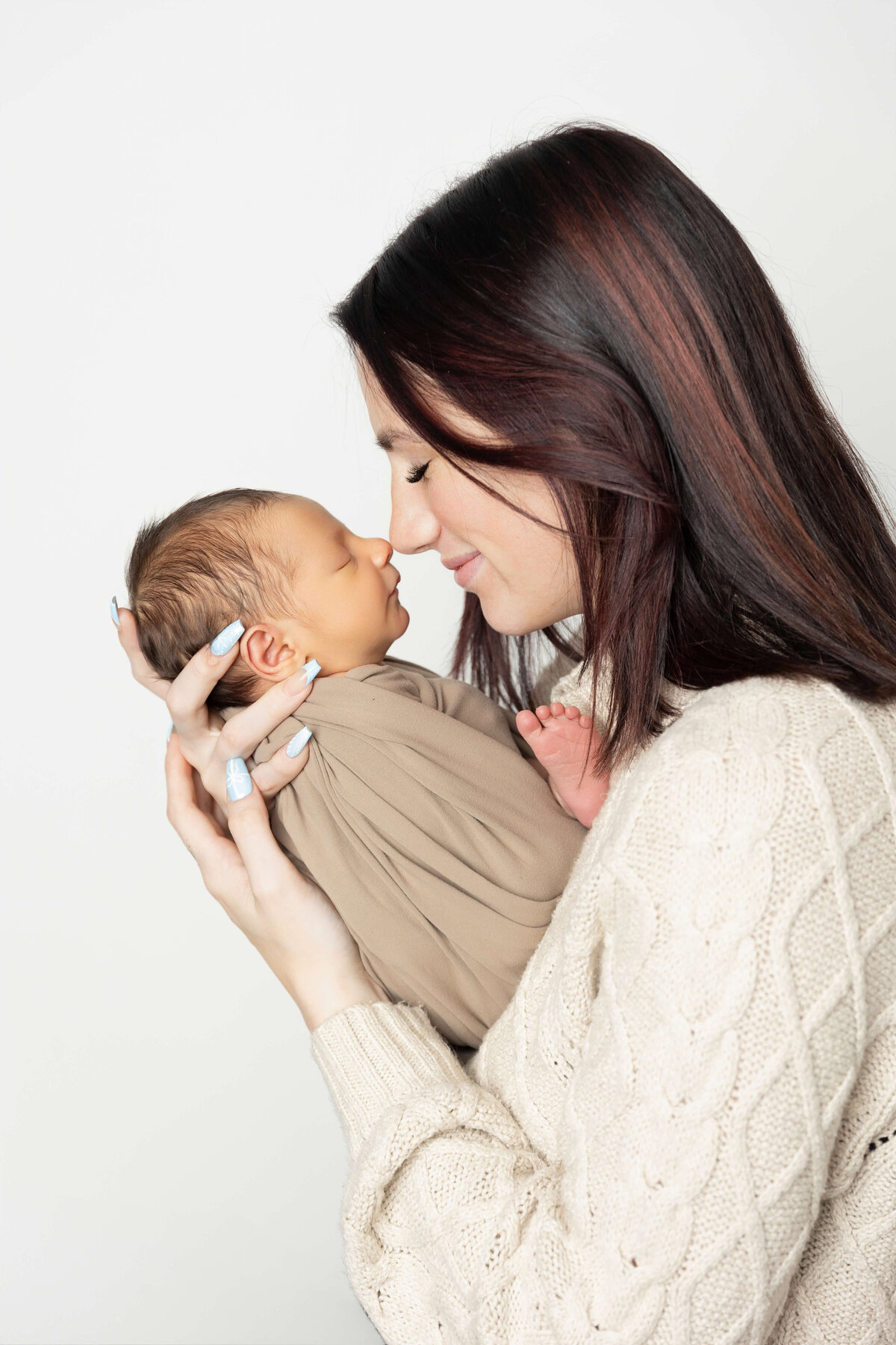 A mother cradles her swaddled newborn in her arms, their faces close together as they share a warm, affectionate moment. The mother has long hair and is wearing a cozy, light-colored sweater. The background is white, creating a soft, tranquil atmosphere.