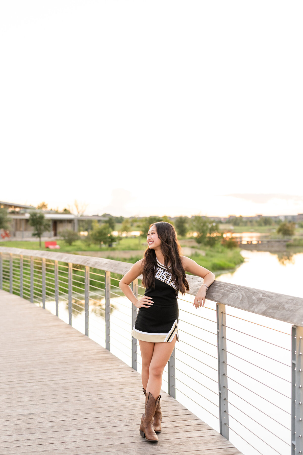 Houston High School senior girl wearing cheerleading outfit and leaning on bridge with hand on hip while smiling off camera at Josey Lake Park
