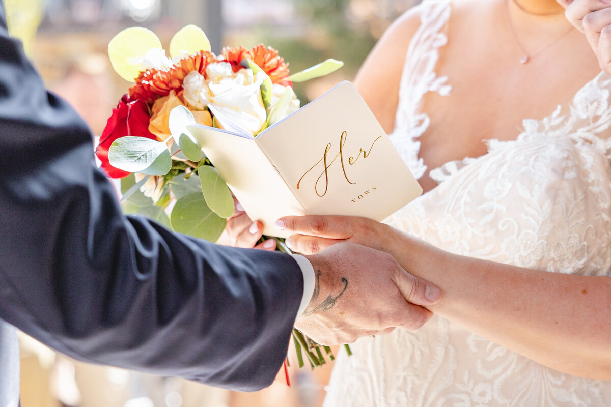 The groom is holding the brides hands as she reads her vows from a book. 