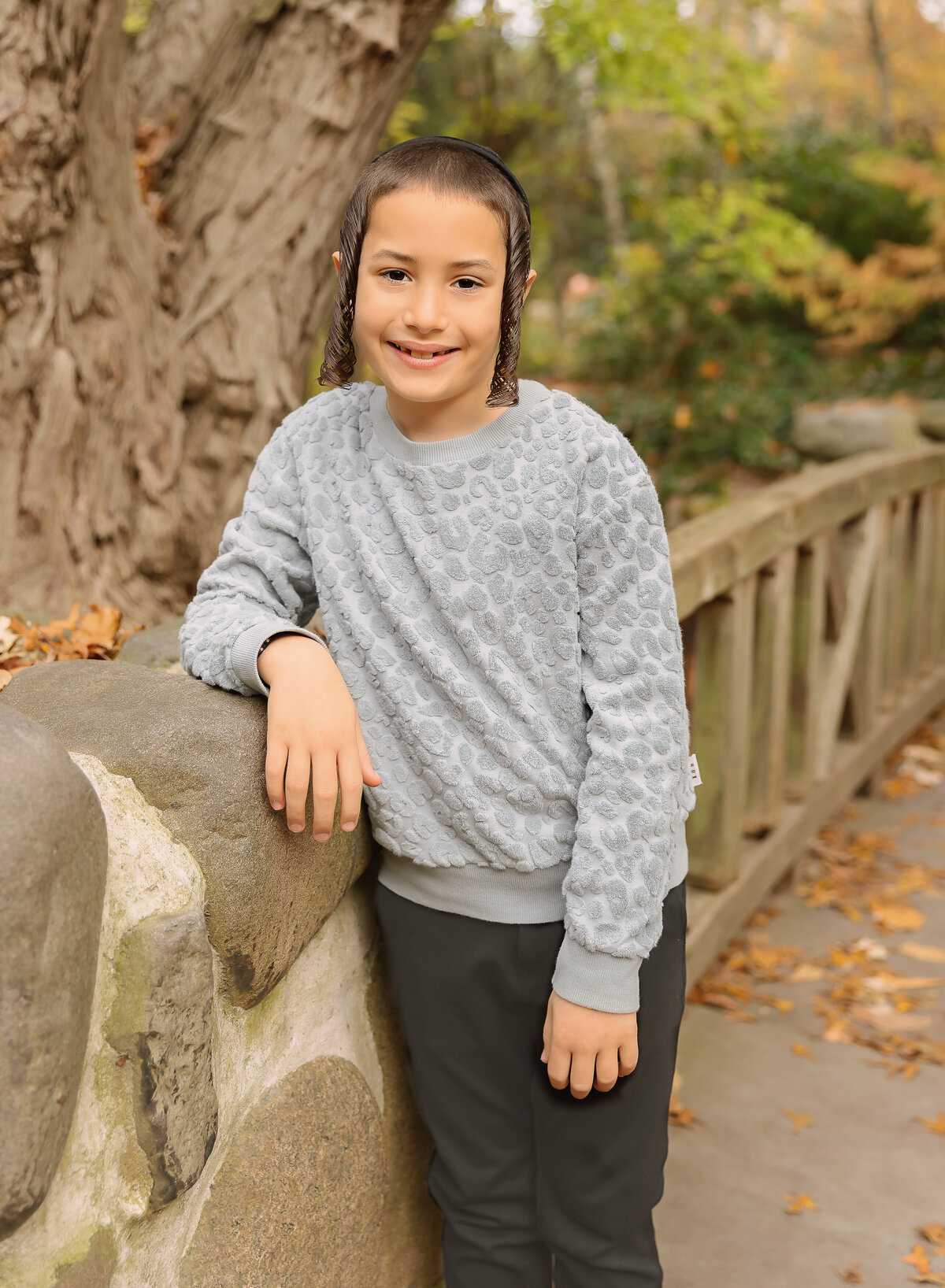 Little boy stands on a stone bridge in brooklyn, ny for a fall familiy photoshoot. His arm is resting on the bridge and smiling at the camera. Captured by Brooklyn family photographer Chaya Bornstein