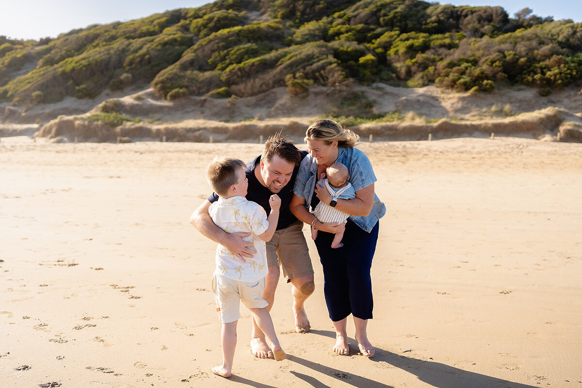 Candid beach family photography in Geelong showing parents playing with their two young children on the sand with coastal cliffs in the background.