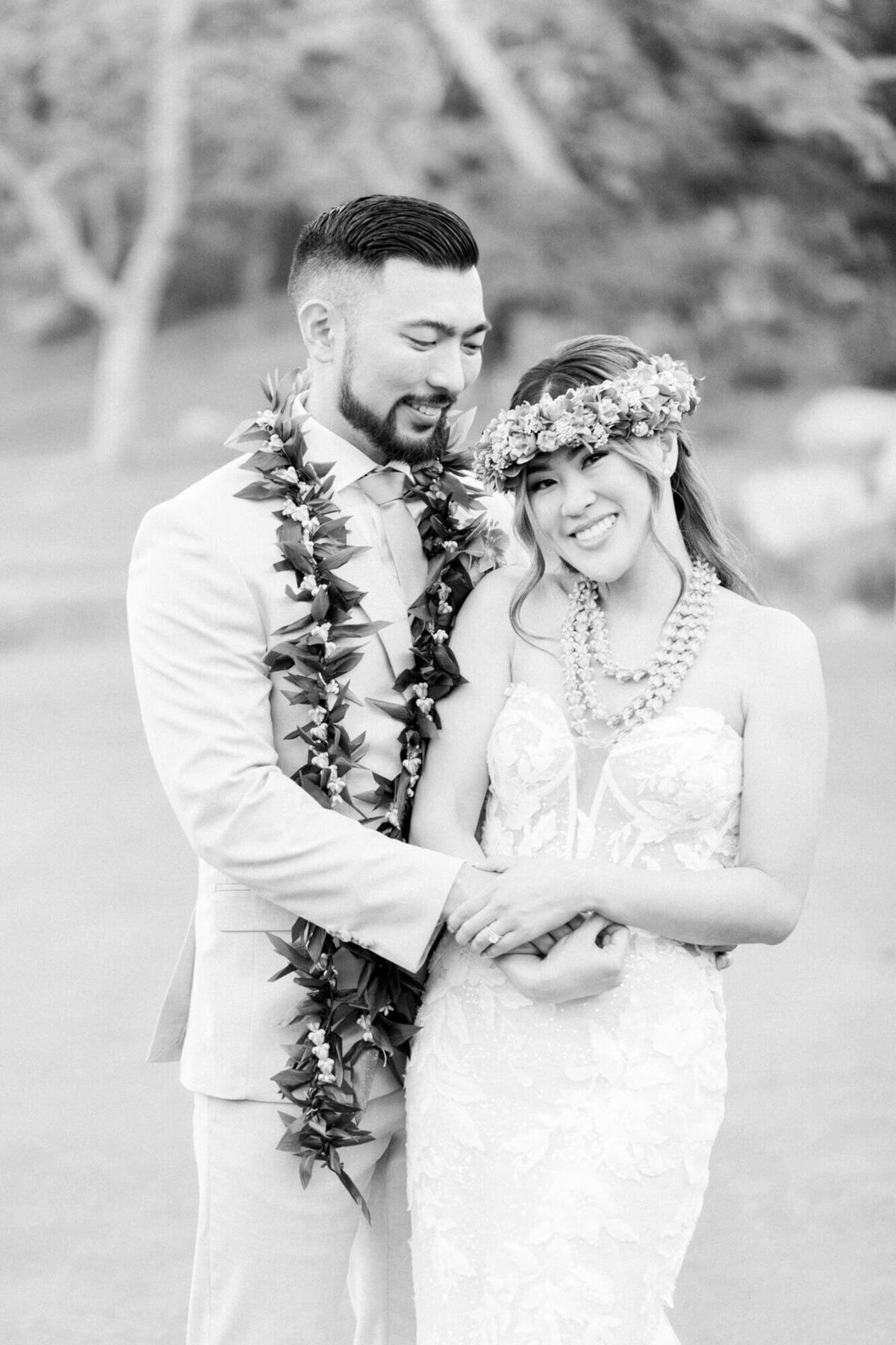 A happy couple in wedding attire embraces outdoors. The bride wears a lei and floral crown, smiling warmly. The groom wears a suit and lei, gazing affectionately.