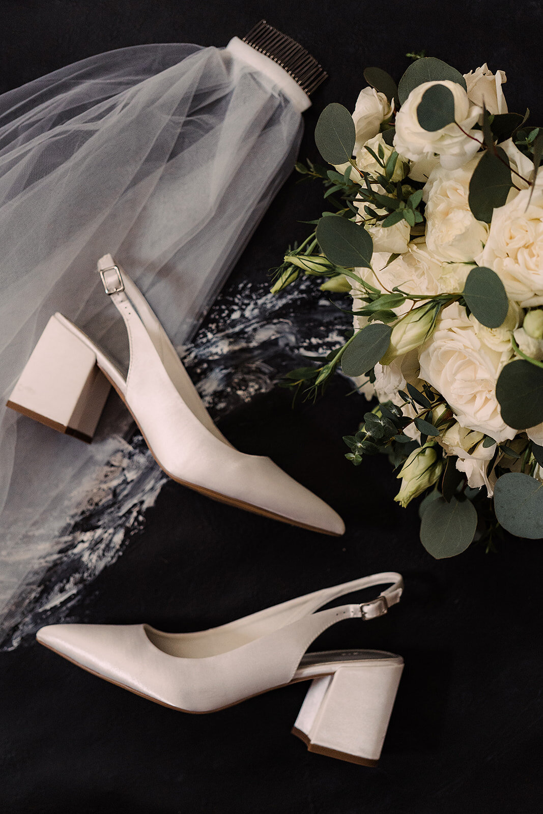 A stylish flatlay of bridal accessories, including white slingback heels, a veil, and a bouquet of white roses with eucalyptus, set against a dark background for a sophisticated touch.