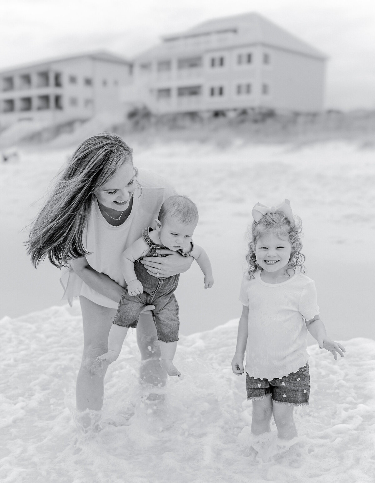 A joyful mother and her two young children play in the waves at Orange Beach, AL. The mother holds her baby securely while her curly-haired daughter, wearing a pink bow, laughs as the foamy ocean water swirls around their feet. Beachfront vacation homes provide a serene coastal backdrop in this timeless black-and-white family portrait