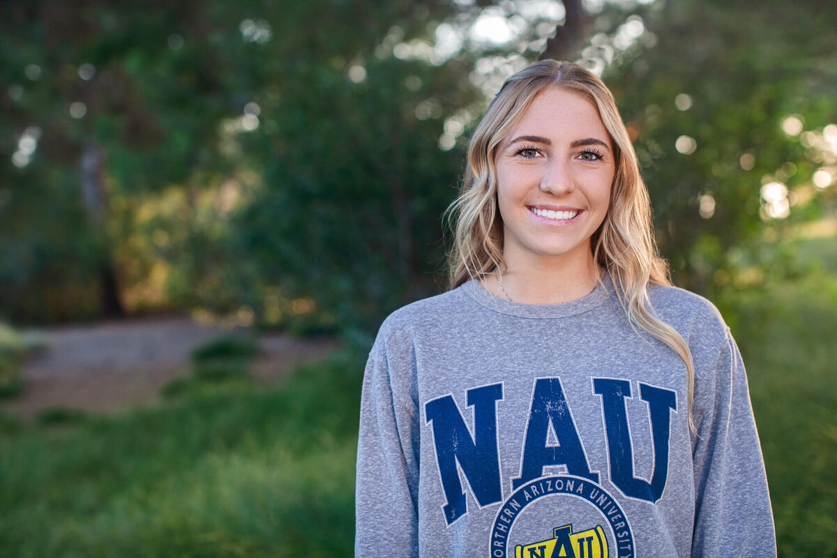 A person stands outdoors, smiling at the camera. They are wearing a gray sweatshirt with NAU printed on it. The background is blurred with green foliage and trees.