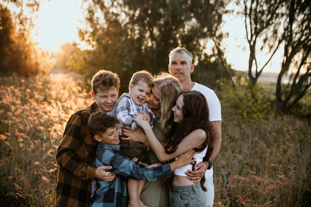 Four siblings hug and tickle the youngest, laughing together in a golden field at sunset.