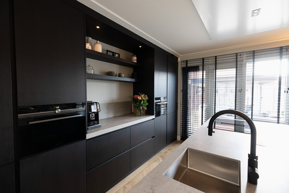 Strakke keukenwand in donker hout met ingebouwde apparatuur en open nissen. De combinatie van gesloten en open elementen geeft deze keuken een moderne, minimalistische look.