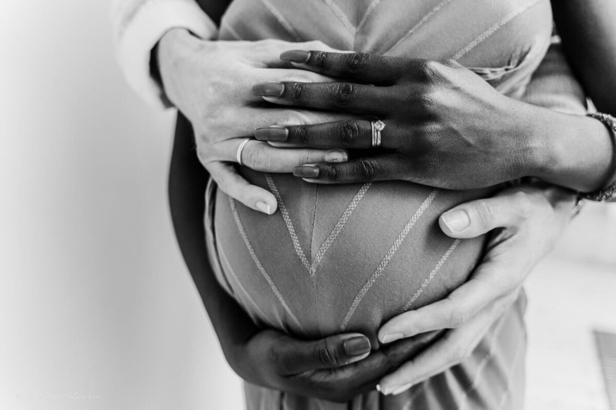 Black-and-white close-up of a pregnant woman in a striped dress with her partner’s hands wrapped lovingly around her baby bump, highlighting the couple’s wedding rings and their shared anticipation.