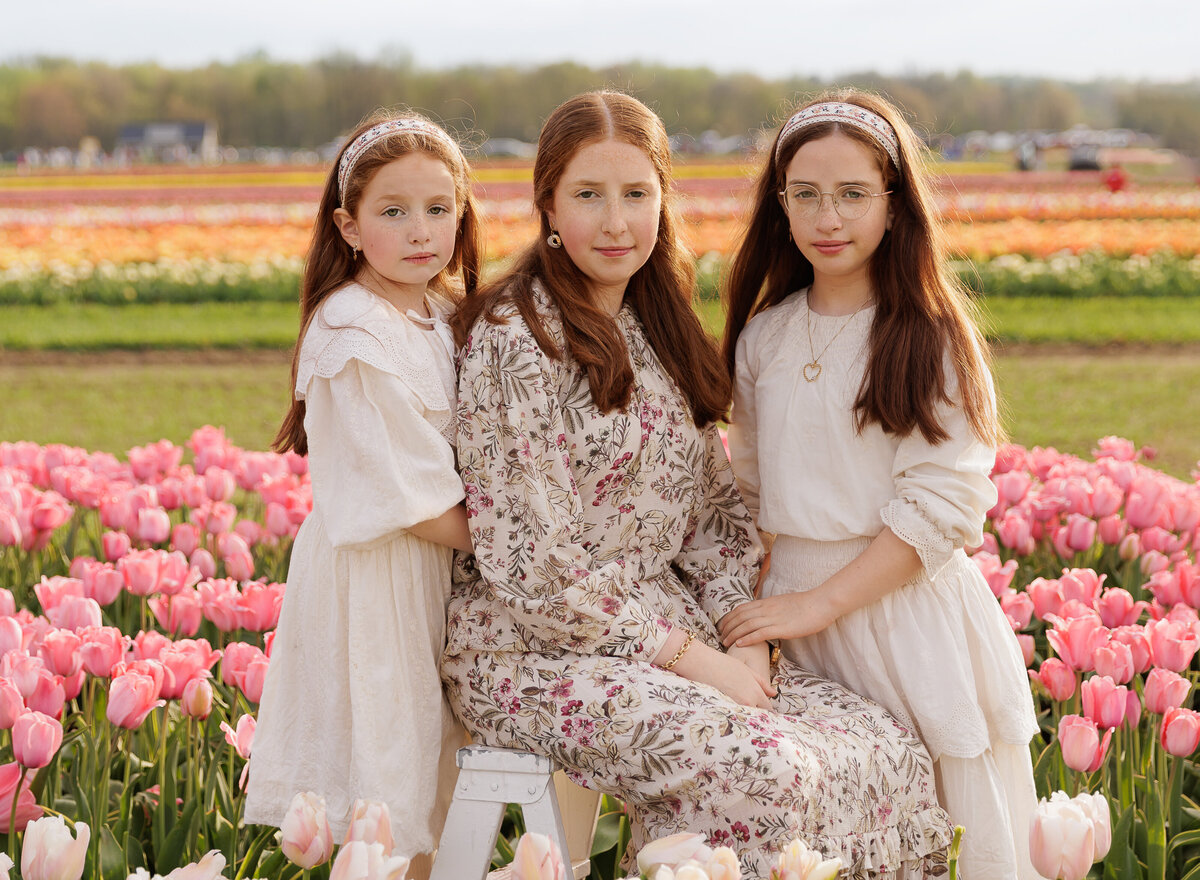 Sisters sit in a field of tulips for a family photoshoot in Brooklyn. All are smiling at the camera. Captured by premier Brooklyn NY family photographer Chaya Bornstein Photography.