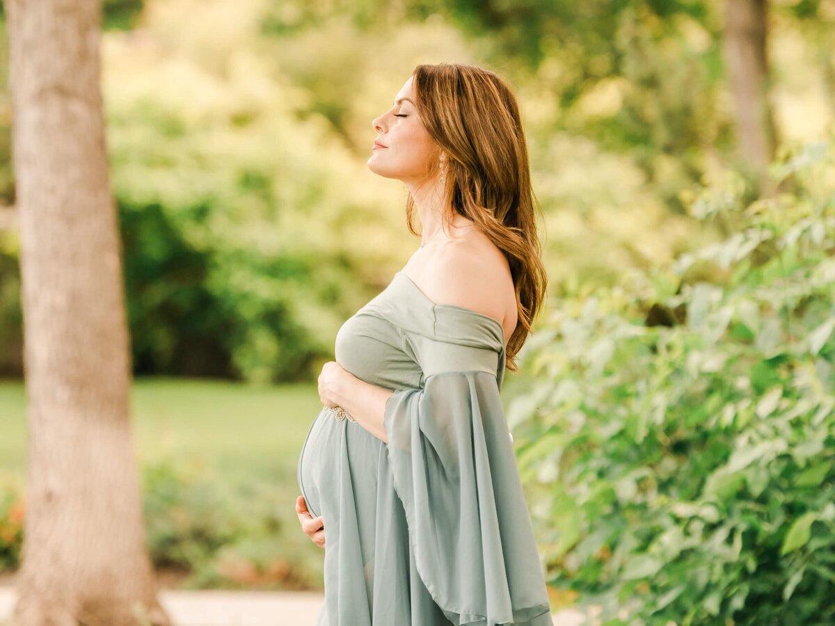 An Edmonton mother holds her belly in an intimate candid moment directed by Cynthia Priest Photography as she breathes and enjoy the golden hour in a nature background in a garden.