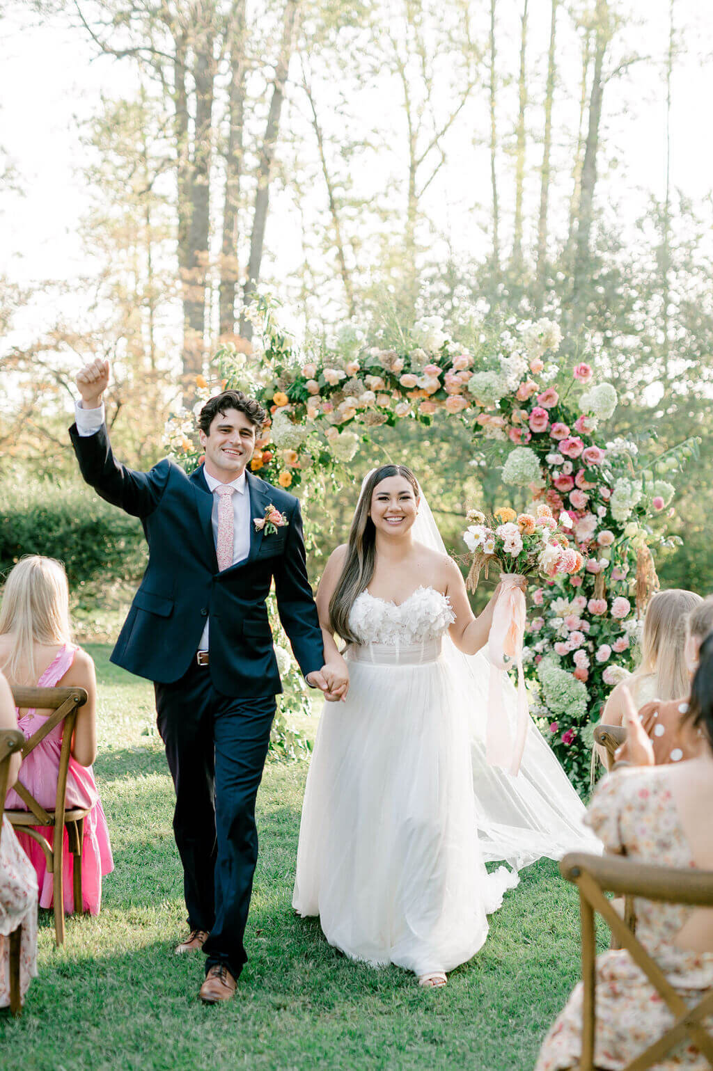 Just married couple walks down the aisle, smiling, under a whimsical floral wedding arch.