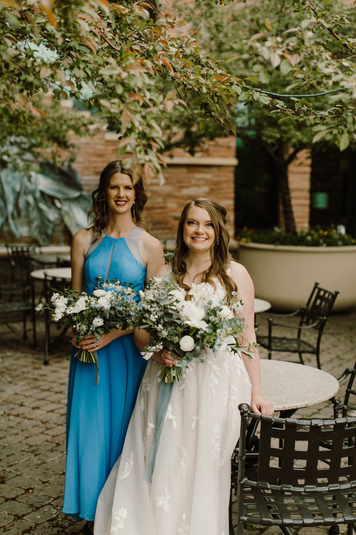 Bride poses with her bridesmaid in a bright blue dress with white and greenery bouquets.