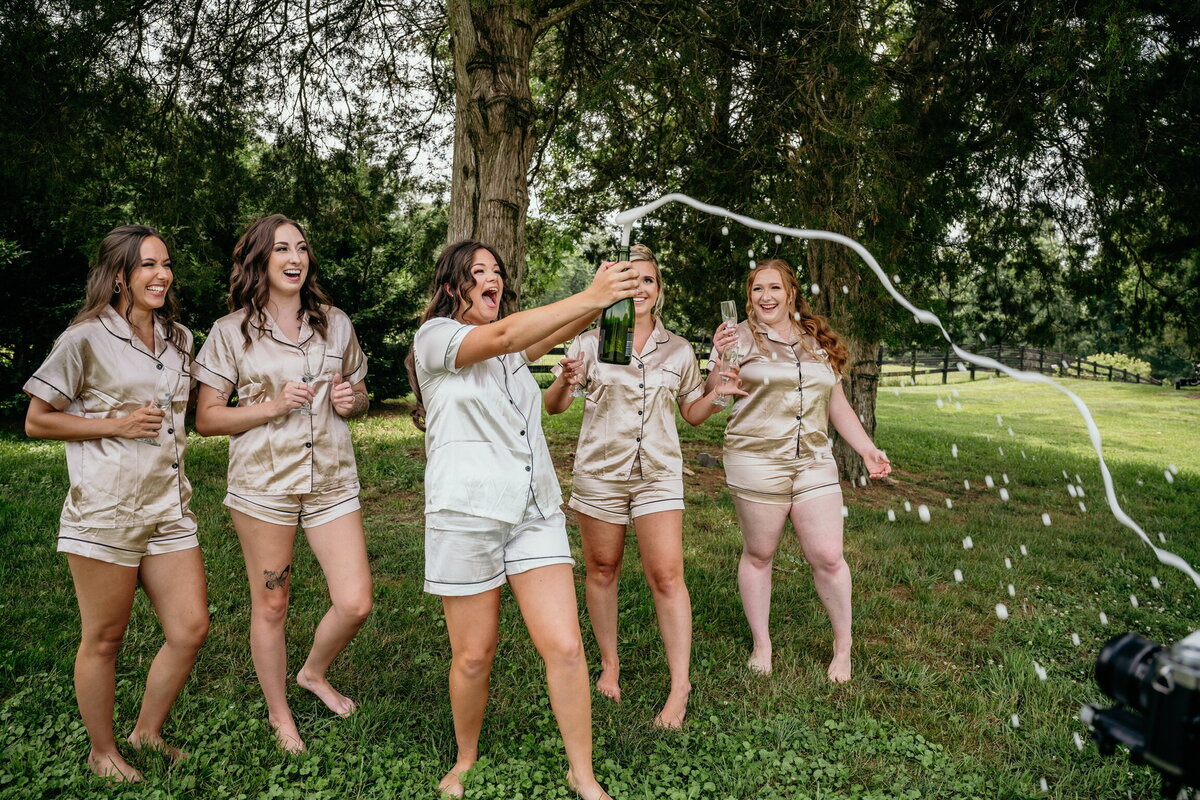 Five women in matching satin pajamas celebrate outdoors, with one popping a champagne bottle, sending a sparkling spray into the air. Laughing on the grass surrounded by trees and an open field, they're enjoying the perfect moment at a rustic wedding venue with ideal event space rentals in Loudoun County.