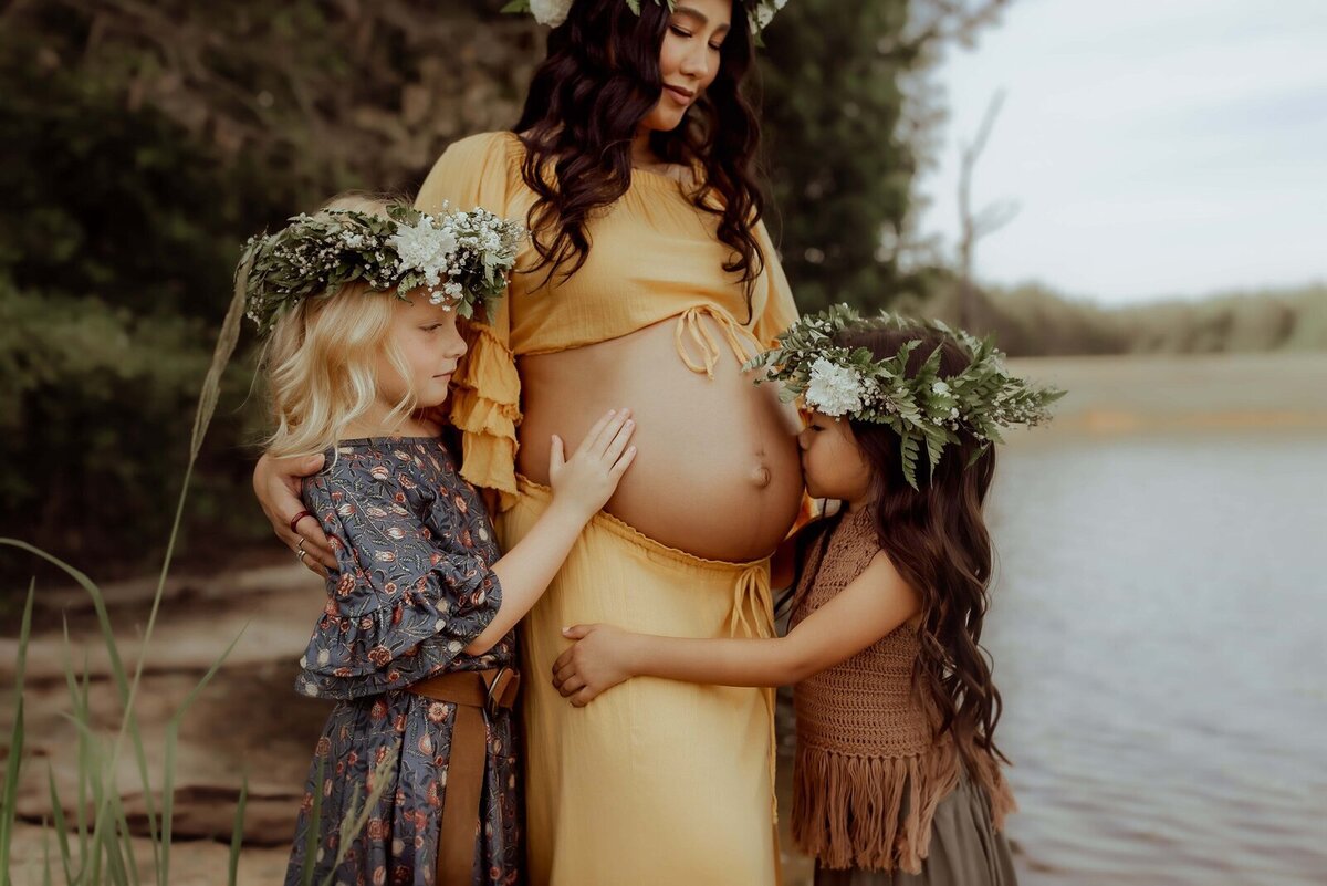 A pregnant woman in a yellow dress, captured beautifully by a Macon maternity photographer, stands by a lake. She holds two children who gently touch her belly, each of them adorned in floral wreaths.