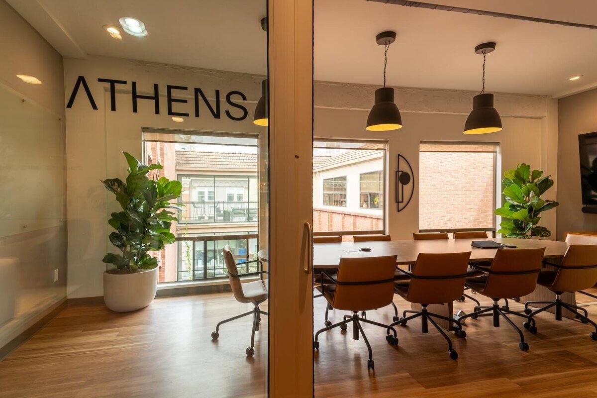 Conference room named “ATHENS” with a long oval meeting table, tan leather chairs, large windows, and tall indoor plants.