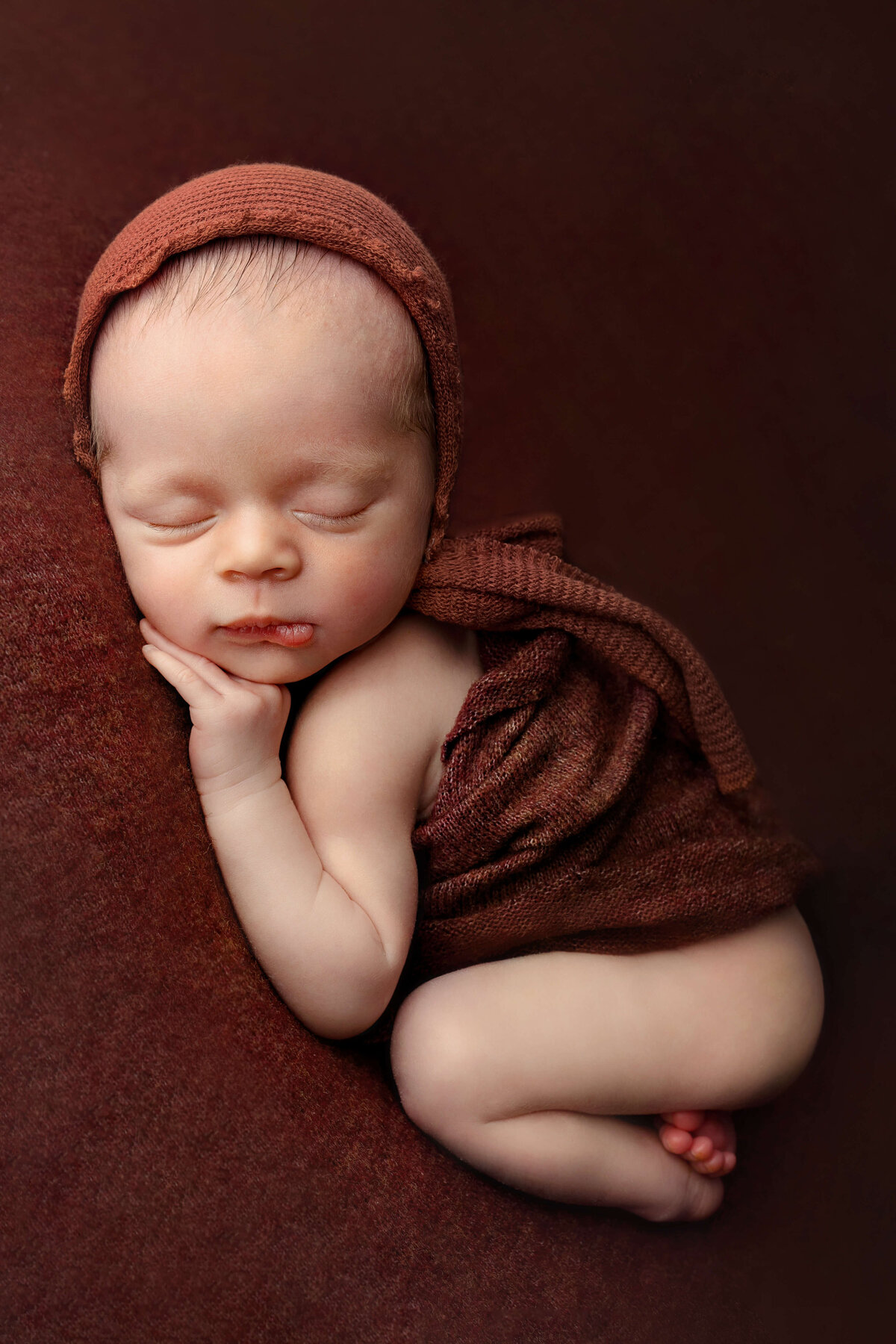 newborn boy laying on his tummy wearing a rust colored bonnet with a rust colored wrap around his tummy on a matching backdrop
