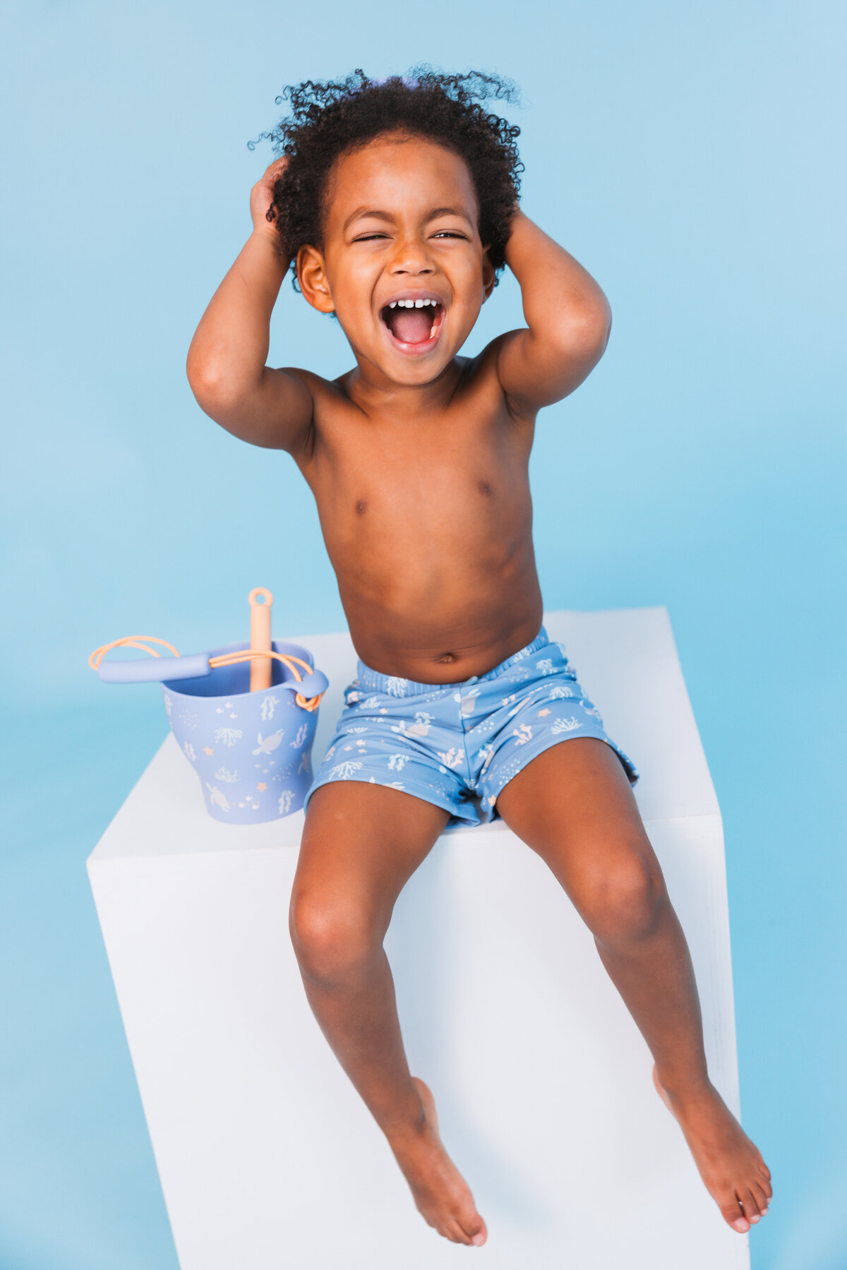 Laughing child in Swim Essentials swim shorts sitting on a white cube, next to a matching blue beach bucket and shovel set.