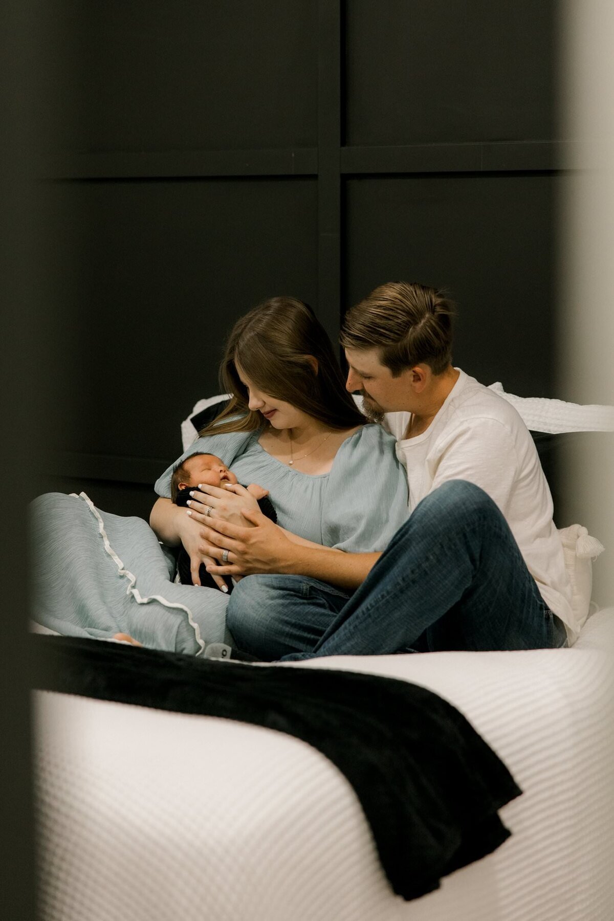 Young couple sitting on their bed, lovingly cradling their newborn baby in a cozy, natural light setting.