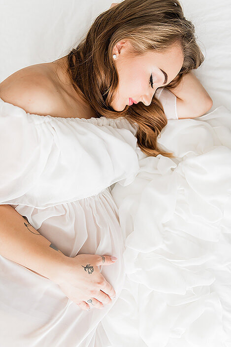 Close-up of a mother-to-be in an off-shoulder ruffled gown, peacefully lying in a soft white setting.