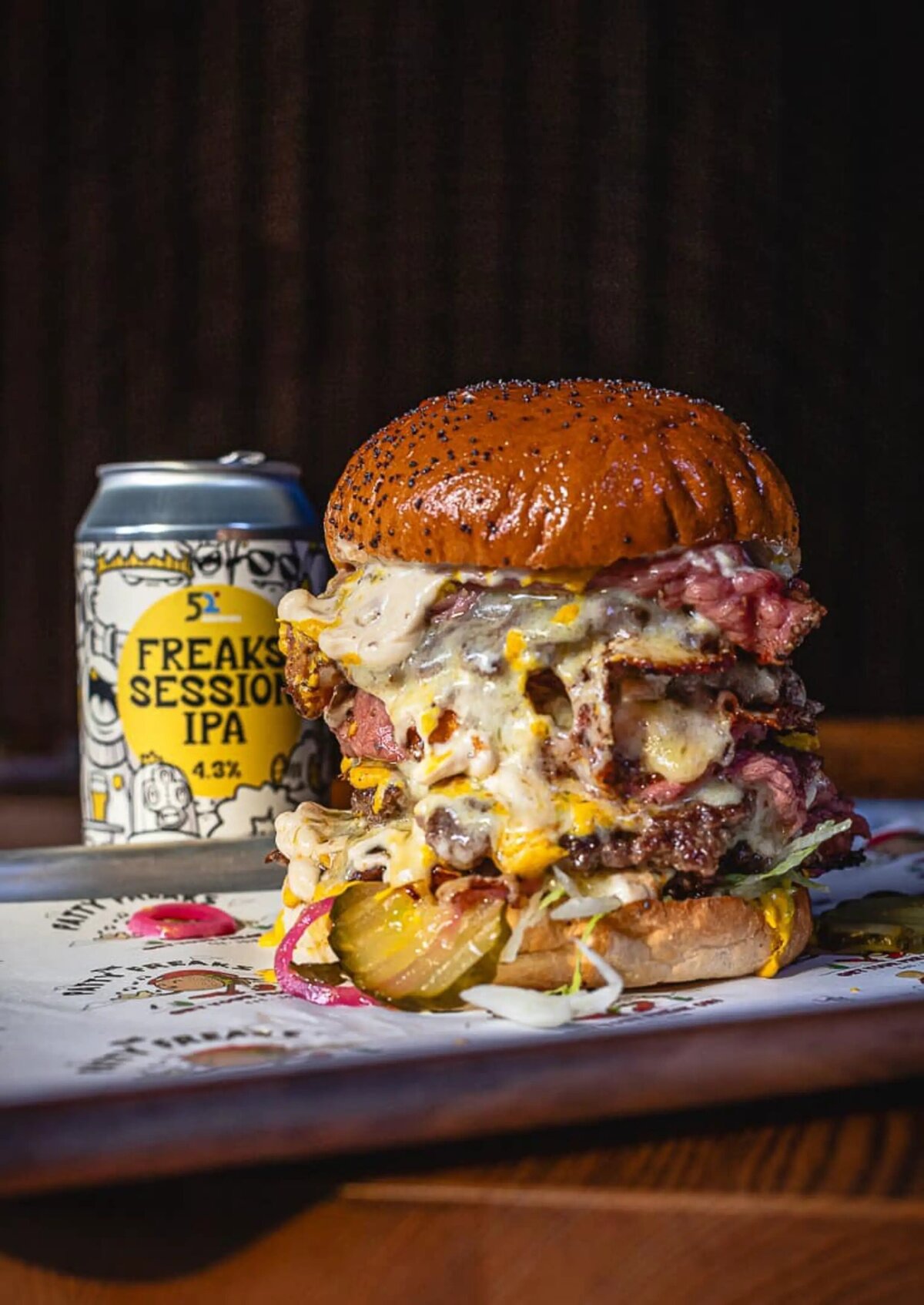 Huge four stacked beef patties oozing out cheese, bacon , lettuce and gherkins. Patty freaks burger on a tray with patty freaks session IPA in the background