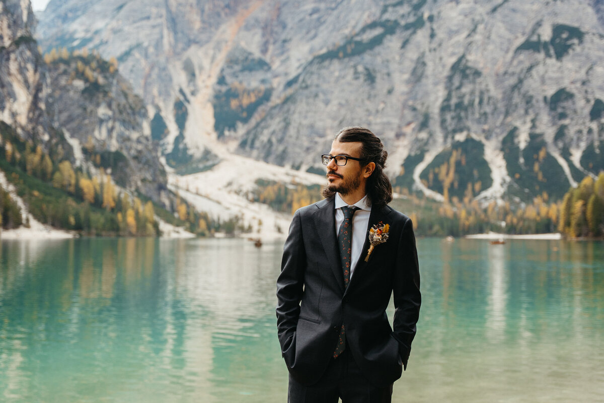 Groom standing by turquoise lake with mountain backdrop
