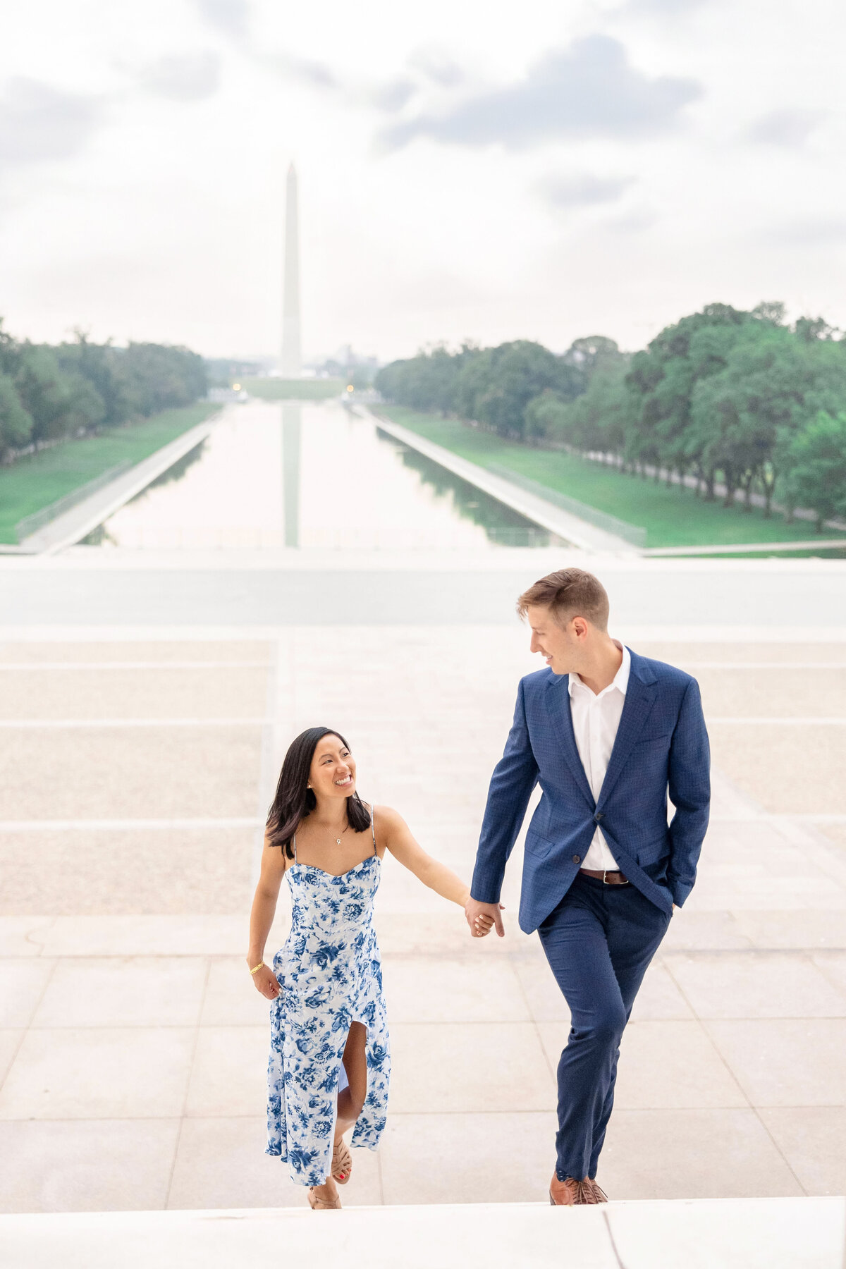 A couple holds hands while walking up white stone steps, with the Washington Monument and Reflecting Pool in the background. The woman is wearing a blue floral dress, and the man is in a blue suit. Trees line the sides.