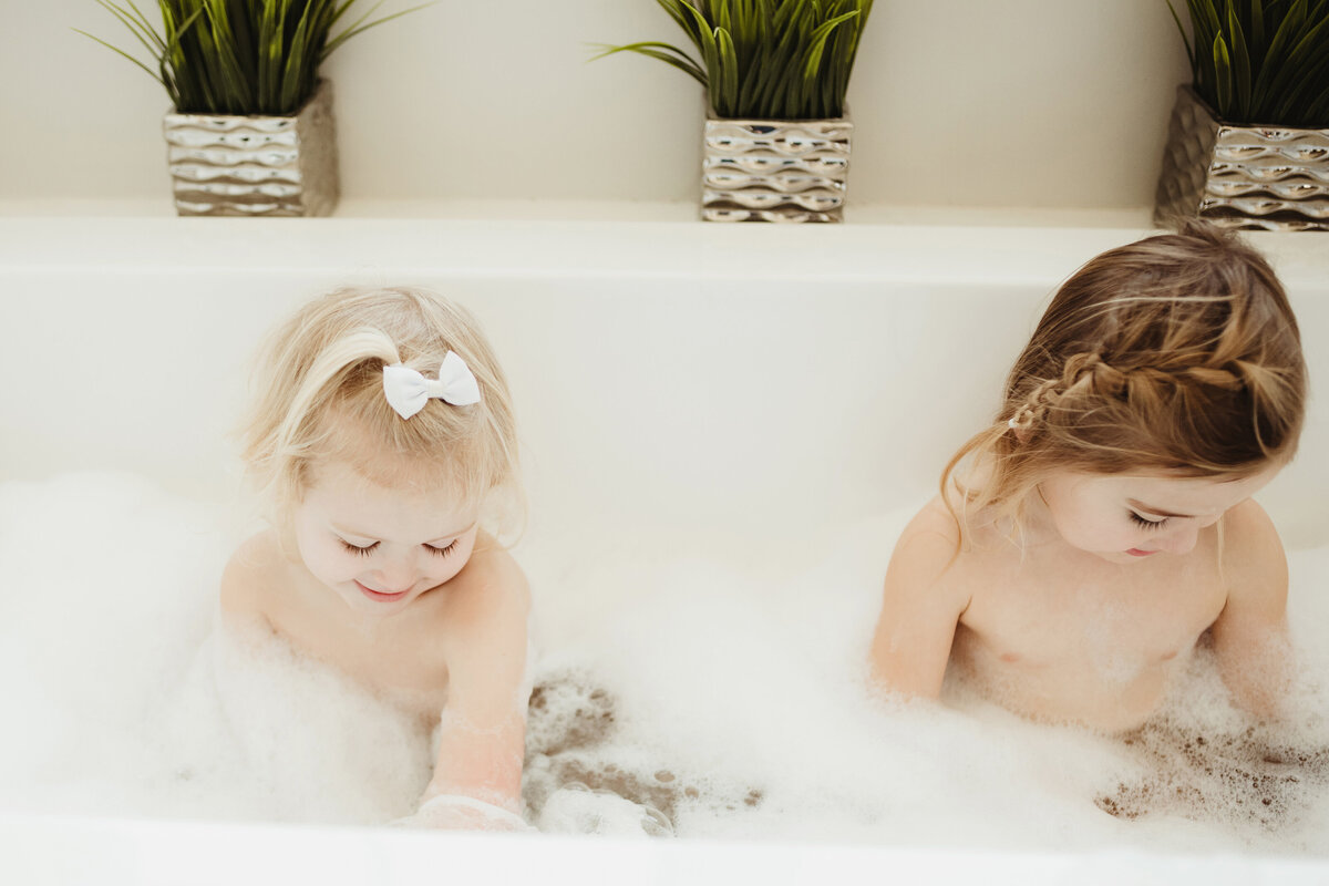 Two toddler girls are playing with barbies in the bubble bath.