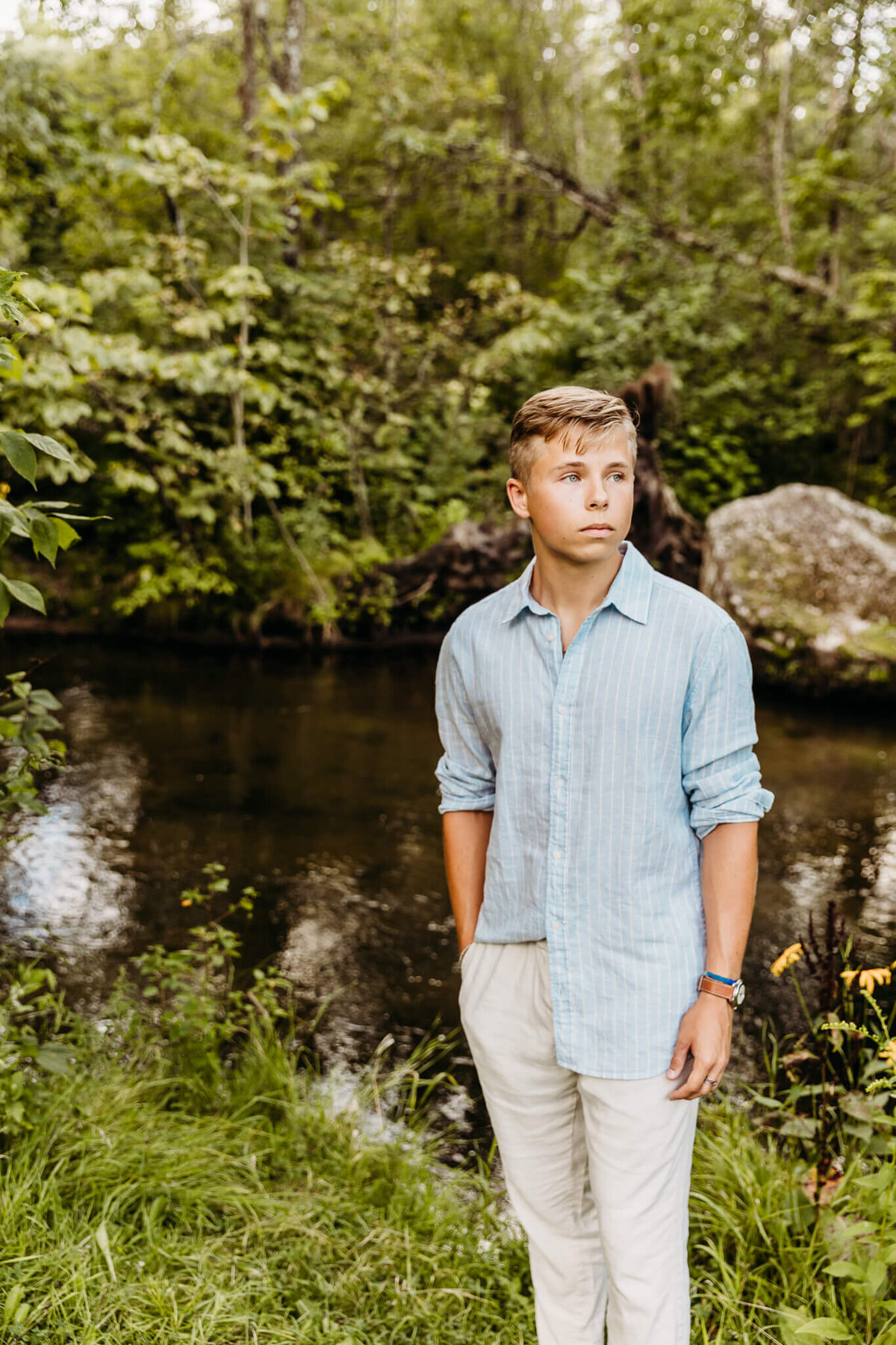 senior boy in white pants and blue shirt standing next to a creek for his senior portraits