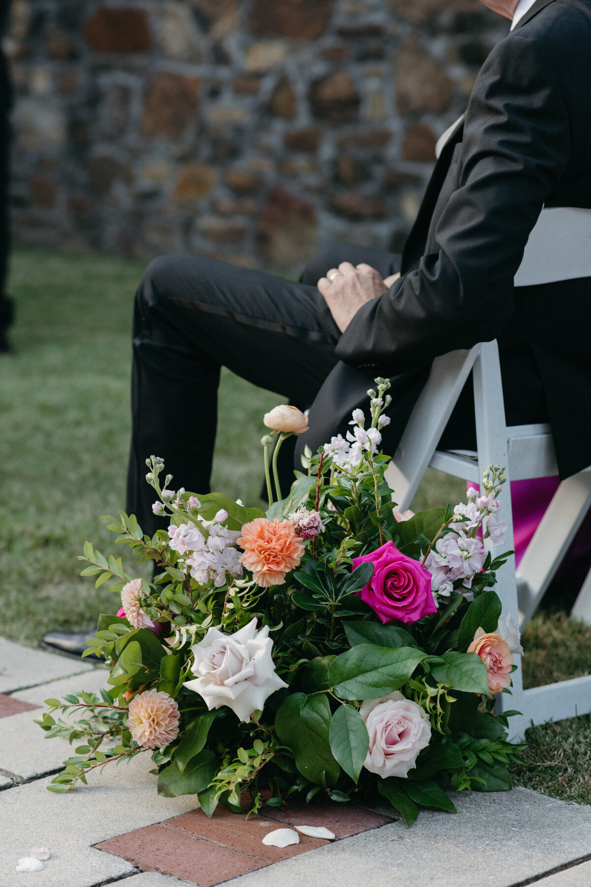 Colorful ceremony ground floral arrangement with roses, dahlias, stock, and lush greenery placed at the aisle entrance, creating a vibrant garden-inspired look for an outdoor wedding.
